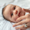 A baby lies on a white blanket with a hand gently touching their face. The baby has wide eyes and a small amount of Baby Barrier Balm Allergen & Skin Irritant Free is visible on their forehead. The baby is looking directly at the camera, safe in the knowledge that the product is both skin irritant-free and allergen-free.