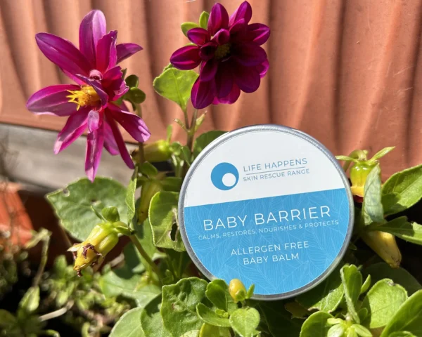 A round container labeled "Baby Barrier Baby Balm" with a blue and white design is placed among vibrant purple and yellow flowers. The container is part of the "Life Happens Skin Rescue Range." The sunlit scene, set outdoors, highlights the natural beauty of this baby barrier balm.