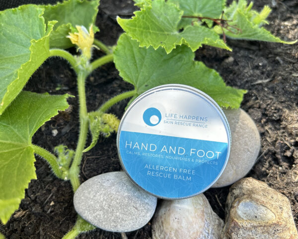 A small tin of "Hand and Foot Rescue Balm" with a blue label lies on top of several stones in a garden. Cucumber leaves and a yellow flower surround it.